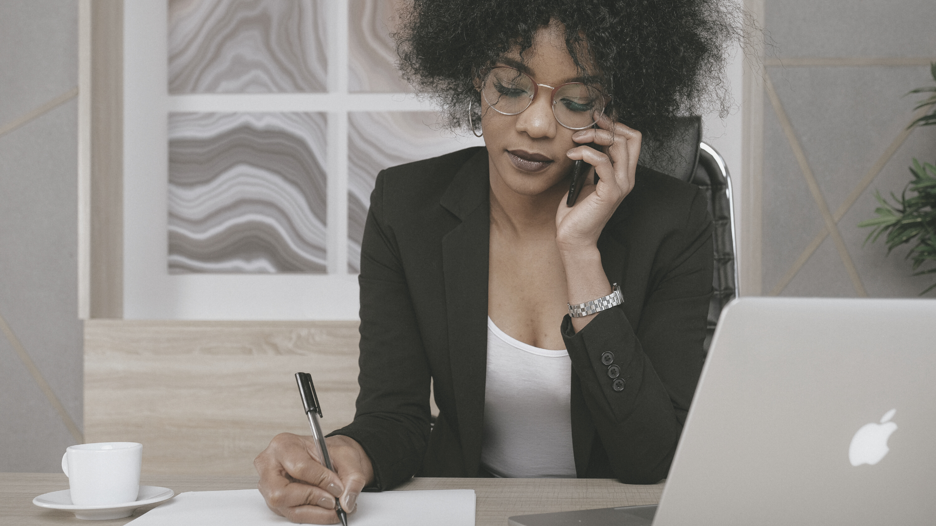 black woman wearing suit sat at desk on phone writing